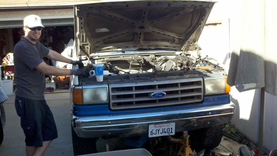 Man working on engine of a blue Ford F-Series truck with hood open. California license plate 6JY401 visible