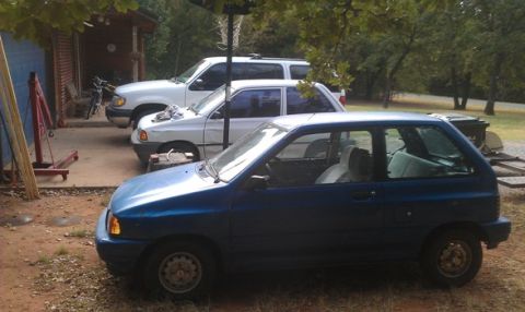 1991 Ford Festiva - Three vehicles are parked in a driveway next to a building and trees.