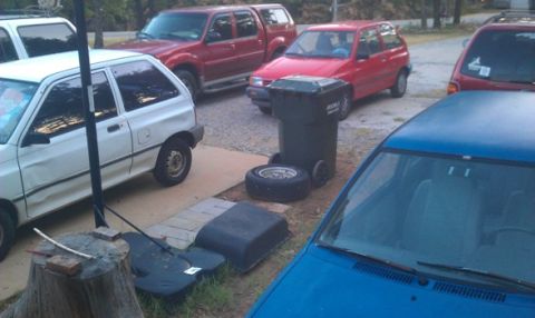 1991 Ford Festiva - A white hatchback is seen parked in a driveway alongside several other vehicles and a trash can.