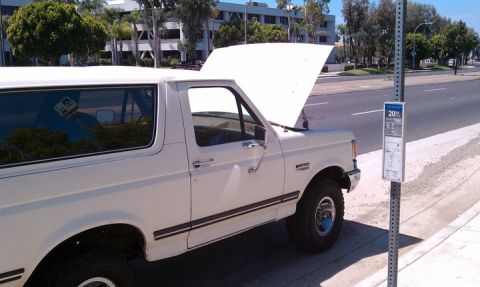 1990 Ford Bronco - A white 1990 Ford Bronco is parked by the side of the road with its hood up, near a bus stop sign.