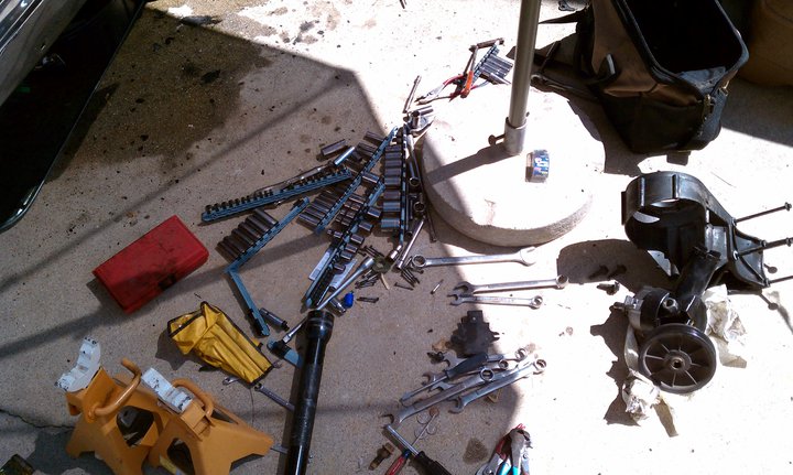 Mechanic's tools, including socket sets and wrenches, scattered on concrete next to jack stands and a toolbox