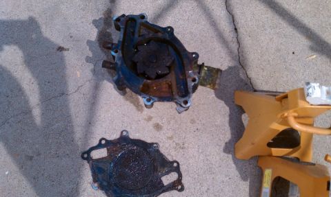 1990 Ford Bronco - Water pump and gasket removed from a vehicle, resting on concrete with a jack stand nearby.