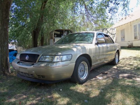 2000 Mercury Grand Marquis - This gold sedan is parked in a grassy area next to a house and some trees. The front of the car is visible.
