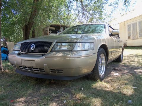 2000 Mercury Grand Marquis - A gold 2000 Mercury Grand Marquis is seen from a three-quarter front view, parked outdoors with a house in the background.