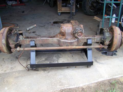 1982 Ford F-100 - Rusty solid front drive axle assembly removed from a truck and set on a low black steel dolly on a garage floor, showing the differential h…