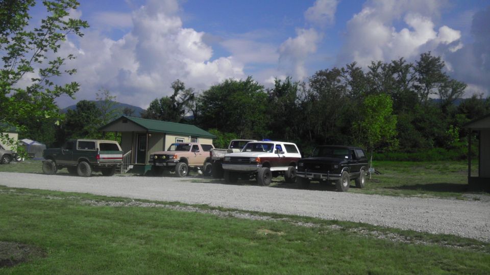 Several SUVs parked on a gravel lot in front of a small building with trees and mountains in the background
