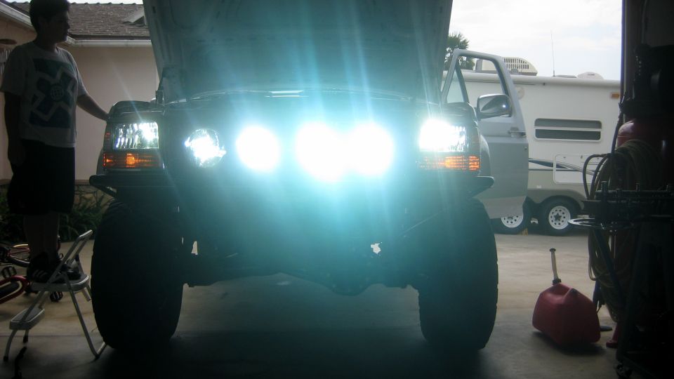 Front view of a 1996 Ford Bronco with bright headlights on, a person stands to the left
