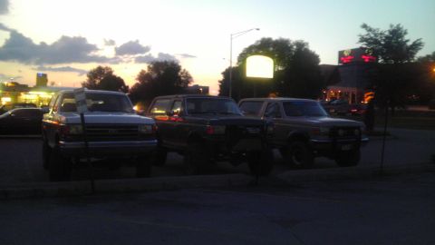 1989 Ford Bronco - Three Ford Bronco SUVs are parked in a lot during twilight.