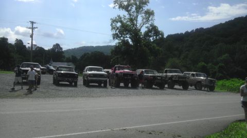 1989 Ford Bronco - A group of off-road vehicles are parked in a lot on a sunny day, with a wooded hillside behind them.