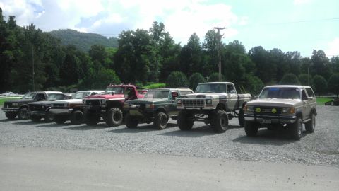 1989 Ford Bronco - Several modified trucks and a tan Ford Bronco are parked in a gravel lot with trees and hills in the background.