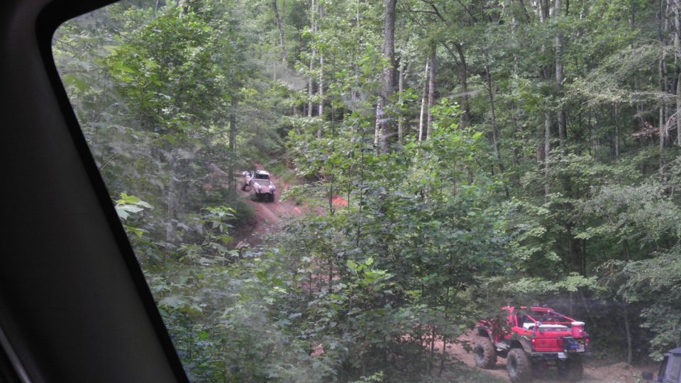 Two off-road vehicles driving on a wooded dirt trail. One is red and in the foreground, the other is lighter colored and further up the trail