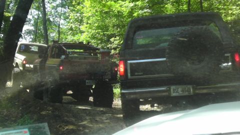 1989 Ford Bronco - Several modified off-road vehicles navigate a wooded trail, kicking up dirt as they go.