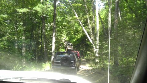 1989 Ford Bronco - Multiple SUVs navigate a wooded trail, kicking up dust on a sunny day.