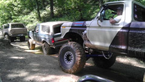 1989 Ford Bronco - Multiple off-road vehicles navigate a muddy, wooded trail, kicking up dirt and water.