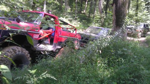 1989 Ford Bronco - Several off-road vehicles are parked in a wooded area, with the closest one being a modified red truck.