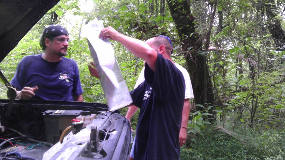 guy with bandana looks at map held by another guy, hood up on vehicle