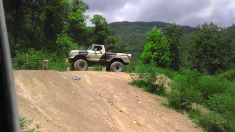 1989 Ford Bronco - Pickup truck modified for off-road use on a dirt track with trees and hills in background.
