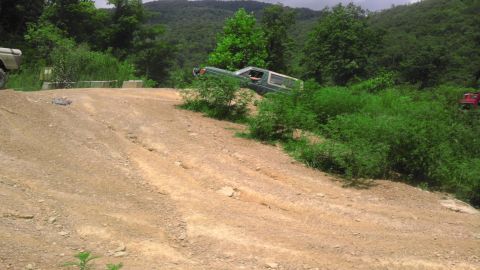 1989 Ford Bronco - An off-road vehicle navigates a steep, dusty trail surrounded by dense green foliage and trees.