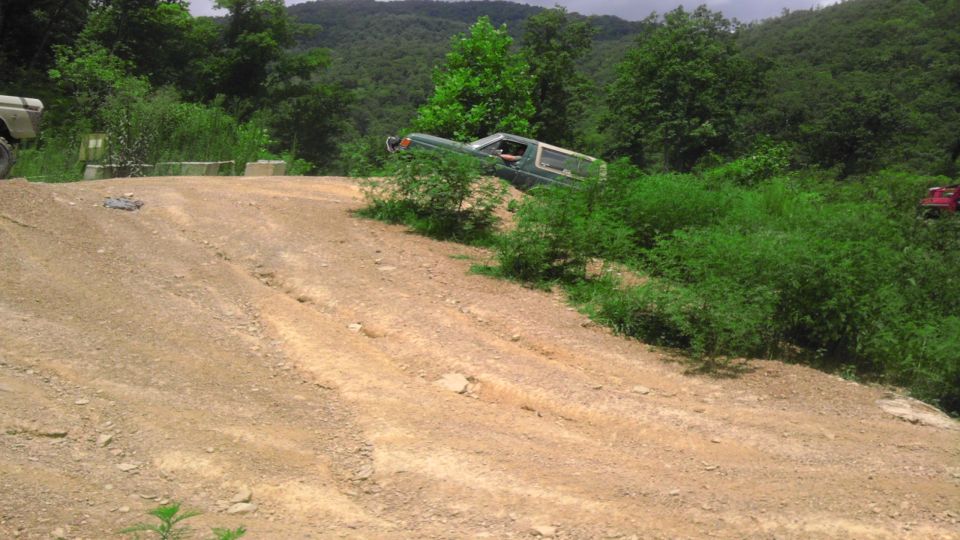 Green SUV tilted on a dirt hill with trees and brush all around. Another white vehicle is on the left and a red vehicle is on the right