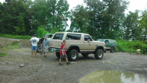 1989 Ford Bronco - A lifted tan 1987 Ford Bronco is parked on a muddy road with other vehicles and people nearby.