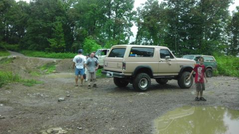 1989 Ford Bronco - Several SUVs, including a tan Ford Bronco, are parked on a dirt road in a wooded area.