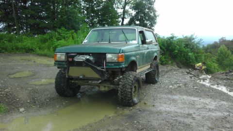 1989 Ford Bronco - This modified SUV is parked on a wet, muddy path with trees and hills in the background.