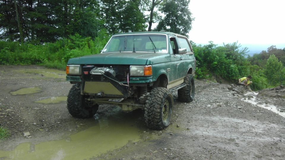Green SUV with large tires on a muddy trail, front grille removed, winch visible