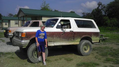 1989 Ford Bronco - Two trucks are parked outside cabins, one is a white and brown Ford Bronco covered in mud.