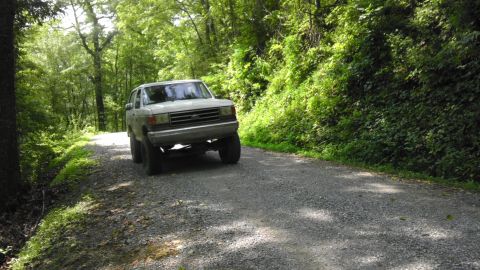 1989 Ford Bronco - A white Ford Bronco is parked on a gravel road surrounded by trees.