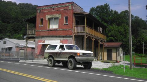 1989 Ford Bronco - A dirty Ford Bronco is parked on the side of the road in front of an old brick building.