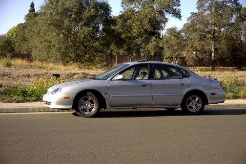1998 Ford Taurus - Side view of a silver sedan parked on the side of a road with trees and vegetation in the background.