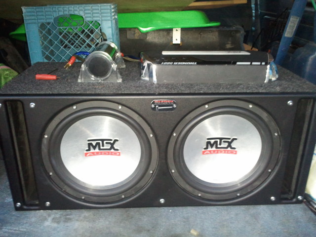 Dual MTX subwoofers and an amplifier in a box, with a blue milk crate and green lid behind them