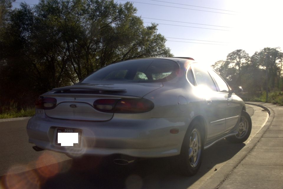 Silver 1998 Ford Taurus sedan, rear 3/4 view, parked on a paved surface with trees and power lines in background