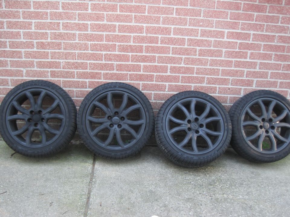 Four black alloy wheels with tires against a brick wall