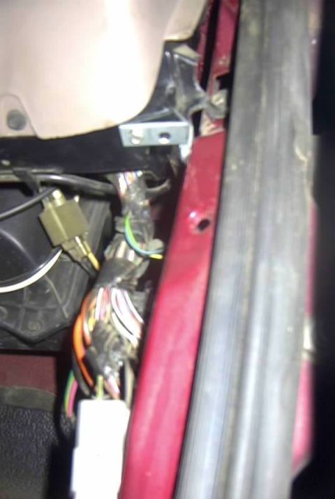 1994 Ford Bronco - Blurry close-up inside a red vehicle's door-jamb and kick-panel area, showing a taped wiring harness with multicolored wires, a white conne…