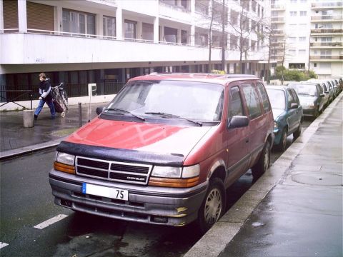 1991 Ford LTD Crown Victoria - A red minivan is parked curbside on a wet street, with several other vehicles lined up behind it.