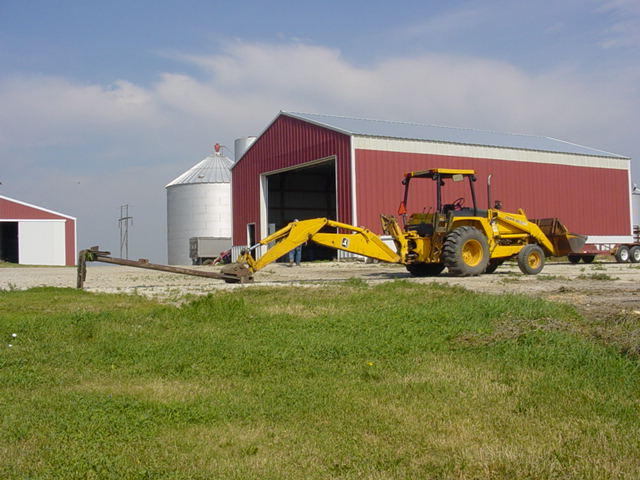 Yellow backhoe loader parked in front of a red barn with silos in the background