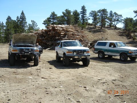 1996 Ford Bronco - Three Ford Broncos are parked in a dirt lot with a large pile of logs behind them under a clear blue sky.