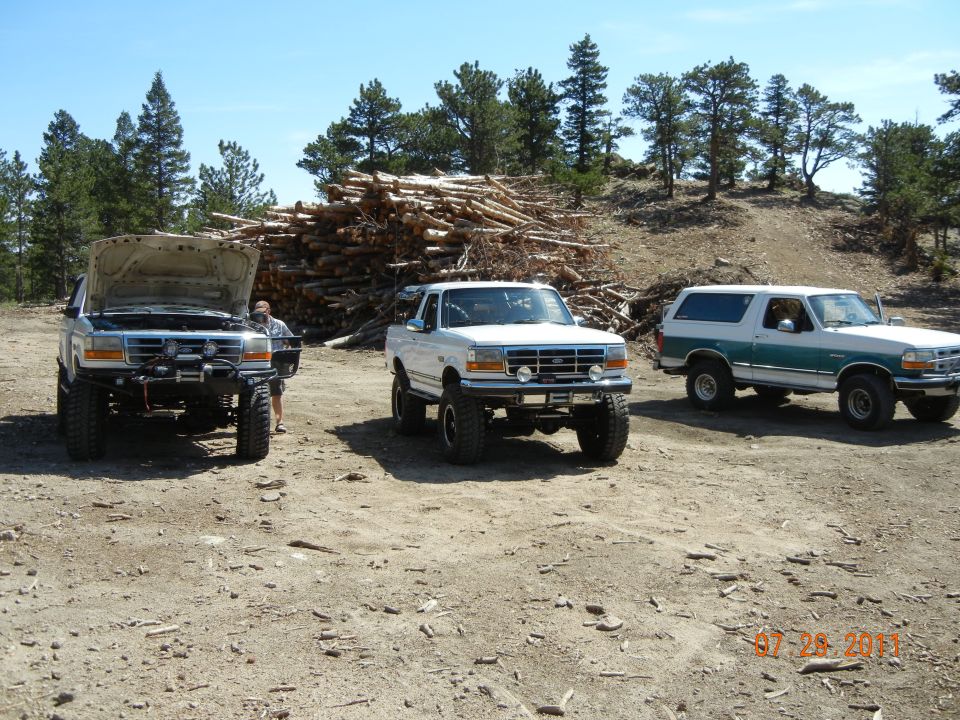 Three lifted Ford Broncos parked in a dirt lot. One has its hood open. A pile of logs is in the background