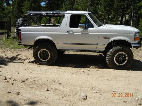 1996 Ford Bronco - A white Ford Bronco is shown on a dirt road with trees in the background. The vehicle has been modified for off-road use.