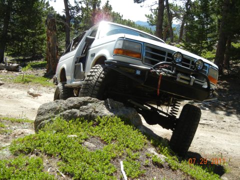 1996 Ford Bronco - This modified SUV is navigating a rocky obstacle on an off-road trail, with trees and mountains in the background.