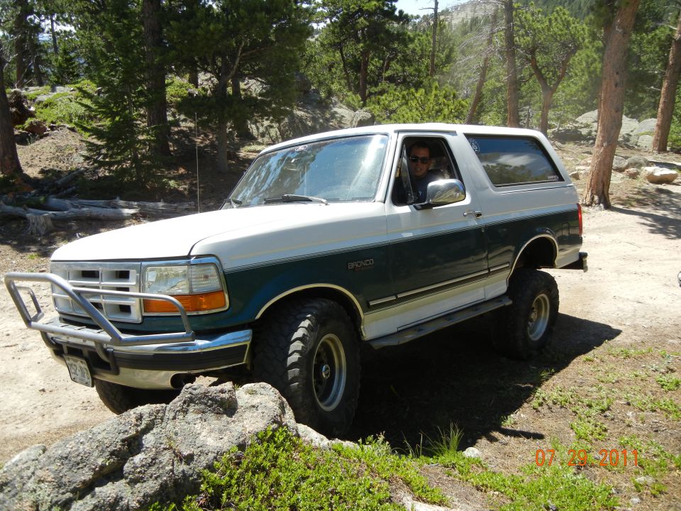Two-tone green and white Ford Bronco on a dirt trail with trees and rocks