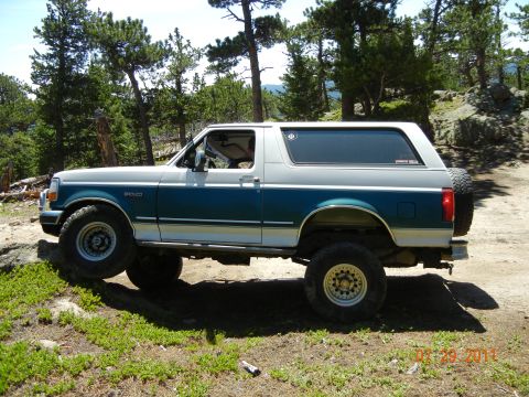 1996 Ford Bronco - A 1996 Ford Bronco is shown in a side profile, parked on a dirt path surrounded by trees and rocky terrain.
