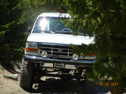 1996 Ford Bronco - A white 1996 Ford Bronco is seen from the front, modified for off-road use with large tires and a winch, partially obscured by trees.