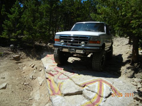 1996 Ford Bronco - A white Ford Bronco navigates a rocky, off-road path with trees in the background.