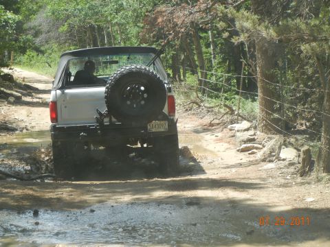 1996 Ford Bronco - A Ford Bronco navigates a muddy trail surrounded by trees, with a spare tire mounted on the back.