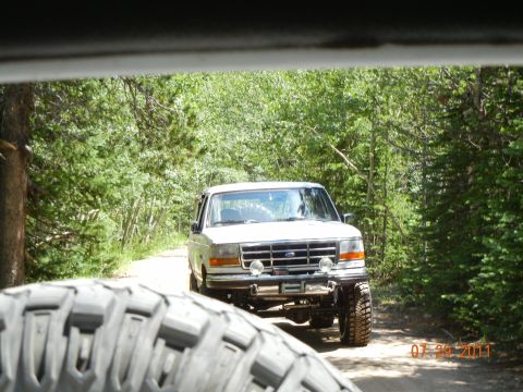 1996 Ford Bronco - White 1996 Ford Bronco drives on a dirt road surrounded by trees.