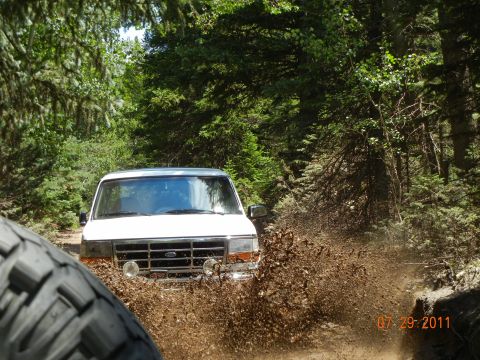 1996 Ford Bronco - A white Ford Bronco navigates a muddy trail, kicking up a spray of dirt and water.