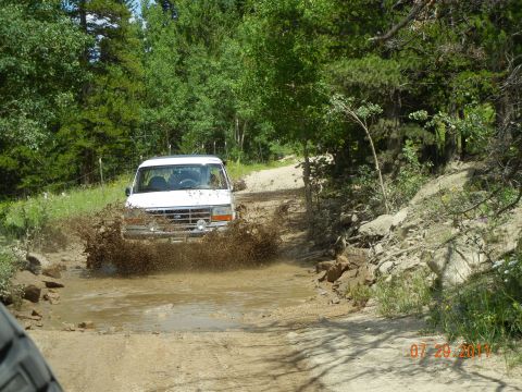 1996 Ford Bronco - A white 1996 Ford Bronco navigates a muddy obstacle on a dirt road, kicking up a large spray.
