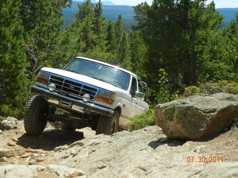 1996 Ford Bronco - A white Ford F-Series truck navigates a rocky trail with trees and mountains in the background.